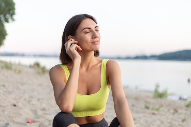 Femme en vêtements de sport au coucher du soleil sur la plage de la ville se reposant après l'entraînement en écoutant de la musique dans des écouteurs sans fil