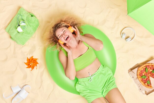 une femme vêtue d'un haut court et d'un short aime écouter de la musique via des écouteurs pose sur une plage de sable seule mange de la pizza se trouve sur une piscine gonflée verte.