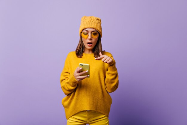 Une femme vigoureuse et stupéfaite en pantalon jaune et pull surdimensionné est surprise par une étrange lettre électronique dans son smartphone. Portrait lumineux du modèle émotionnel contre le mur violet