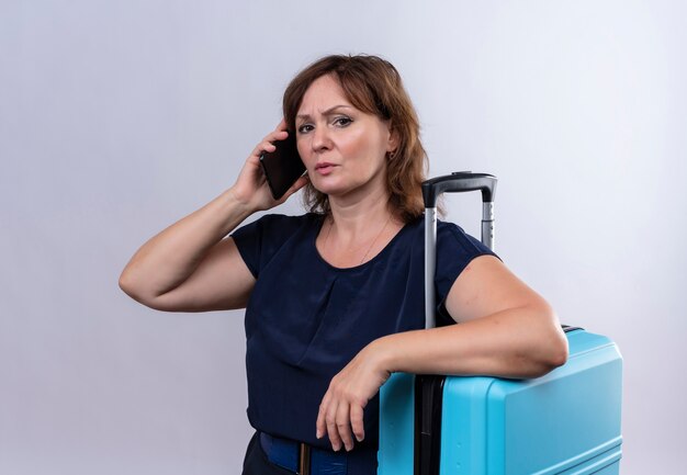 Femme de voyageur d'âge moyen strict parle au téléphone mettant la main sur la valise sur blanc isolé