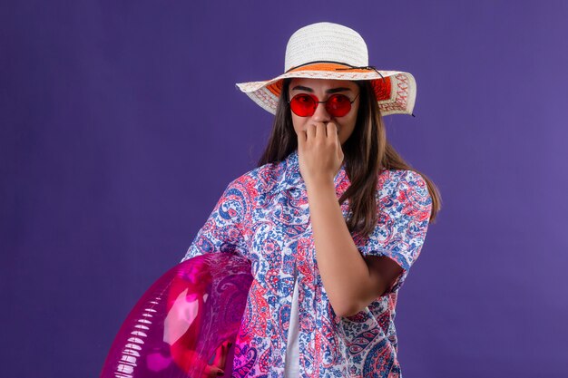 Femme de voyageur portant un chapeau d'été et des lunettes de soleil rouges tenant un anneau gonflable stressé et nerveux de se mordre les ongles debout sur le violet