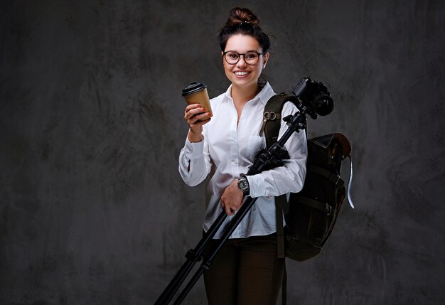 Une femme voyageuse tient un appareil photo numérique et du café à emporter sur fond gris.