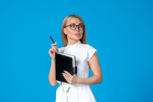 Femme vue de face en robe blanche posant avec un bloc-notes sur un travail bleu