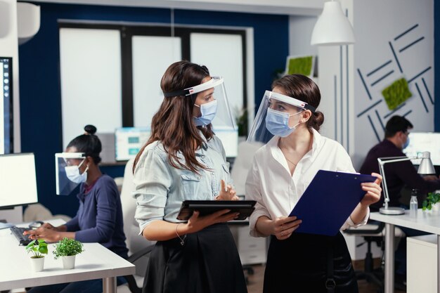 Femmes d'affaires avec des masques de protection contre le coronavirus debout sur le lieu de travail parlant des données financières de l'entreprise détenant une tablette numérique. Équipe commerciale multiethnique travaillant dans le respect de la distance sociale