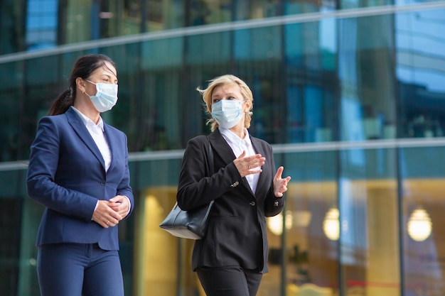 Femmes d'affaires portant des costumes et des masques de bureau, se réunissant et marchant ensemble dans la ville, parler, discuter du projet. Coup moyen. Affaires au cours du concept épidémique