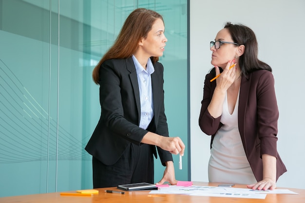 Femmes d'affaires sérieuses discutant des documents statistiques et debout près du bureau dans la salle de conférence