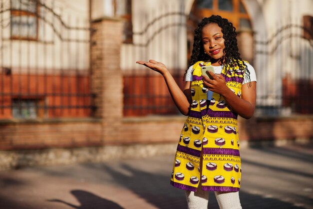 Femmes afro-américaines élégantes en veste jaune posées dans la rue aux beaux jours avec un téléphone portable à portée de main