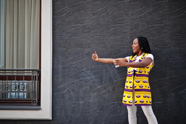 Femmes afro-américaines élégantes en veste jaune posées dans la rue avec une boisson chaude dans une tasse en papier jetable et un téléphone portable à portée de main