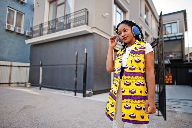 Femmes afro-américaines élégantes en veste jaune posées et écouteurs dans la rue contre un bâtiment moderne