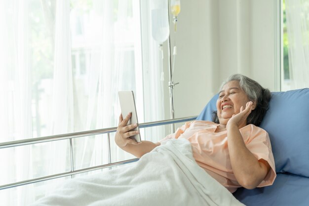 Les femmes âgées dans les patients de lit d'hôpital à l'aide d'appels téléphoniques intelligents à des parents descendants ressentent le bonheur - concept féminin et médical senior