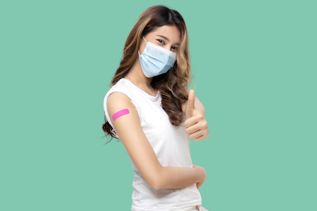 Les femmes asiatiques heureuses avec un masque facial se sentent bien et montrent un bandage sur le bras après avoir reçu le pouce du vaccin Covid19 vers le haut