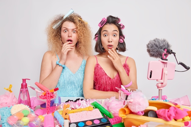 Des femmes diverses et choquées font des robes de coiffure les unes à côté des autres près d'une table pleine de produits cosmétiques enregistrent une vidéo sur la façon de prendre soin de votre apparence pose sur fond blanc