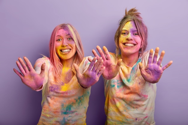 Des Femmes Drôles Célèbrent Les Vacances De Holi, Portent Des Vêtements Blancs Avec Un Spray De Couleur Clairement Visible, Montrent Les Deux Paumes Enduites De Poudre Multicolore, S'amusent, Utilisent Des Colorants Colorés. Arrivée Du Printemps