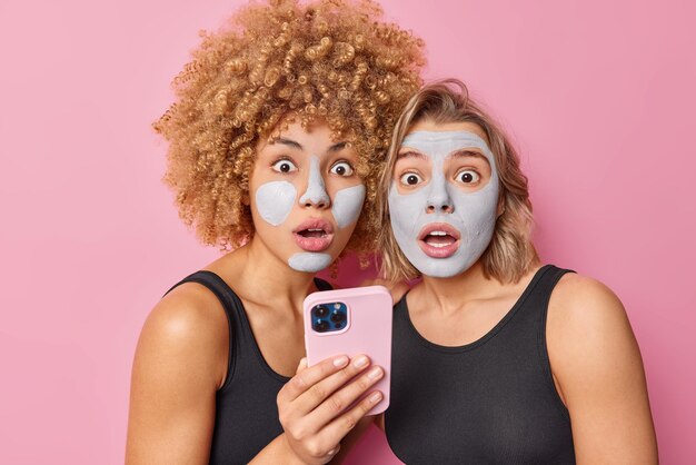 Les femmes effrayées et effrayées réagissent à quelque chose de choquant appliquent des masques d'argile nourrissants sur le visage tient smartphonw subissent des procédures de rajeunissement des soins de la peau posent sur fond rose