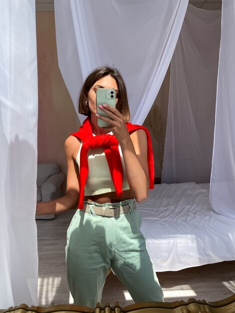 Des femmes élégantes à la maison en pull rouge vif et pantalon vert menthe prennent un selfie photo dans le miroir du téléphone