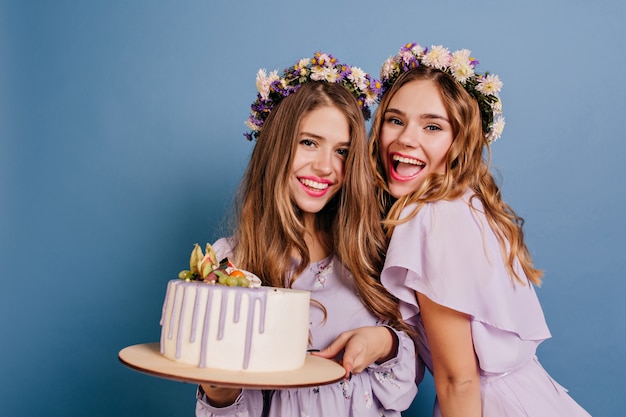 Femmes excitées posant avec un gâteau et riant sur le mur bleu