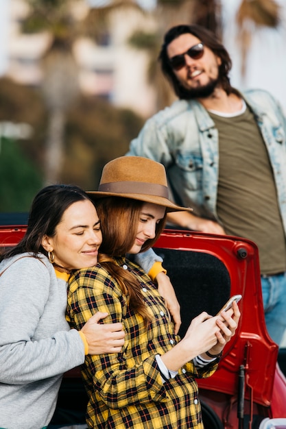 Femmes gaies embrassant dame avec smartphone près de coffre de voiture et homme se penchant de l&#39;automobile