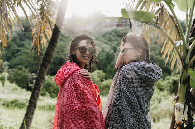 Femmes gaies se promenant dans la forêt en imperméables. Tir en plein air d'amies heureux dans des lunettes de soleil debout sur la jungle.