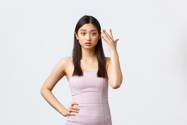 Femmes de luxe, concept de fête et de vacances. Jeune femme asiatique agacée en robe de soirée, pointant vers la tête et l'air irritée, marre d'écouter se plaindre, debout sur fond blanc.