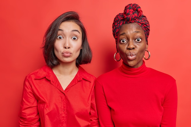 les femmes les meilleures amies se tiennent étroitement les unes aux autres garder les lèvres arrondies attendre le baiser porte des vêtements rouges posent sur le mur lumineux du studio. Modèles féminins de race mixte avec des lèvres plissées. Expressions du visage