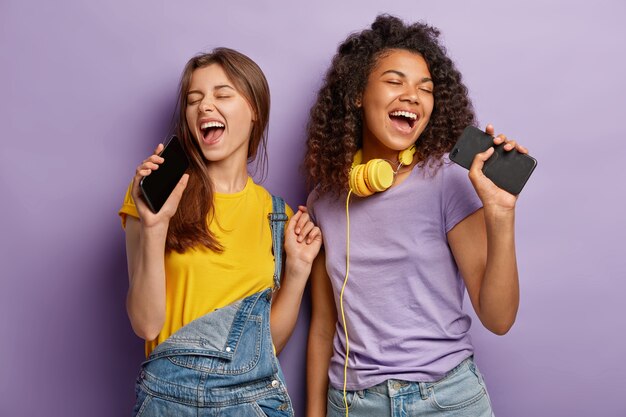 Les femmes métisses optimistes et heureuses chantent la chanson préférée dans les téléphones intelligents, s'amusent et apprécient la musique, gardent les yeux fermés, bougent activement