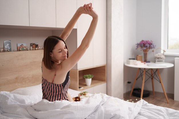 Les femmes profitant d'un café avec des guimauves sur le lit avec un cadeau près d'elle tout en s'étirant après le réveil.