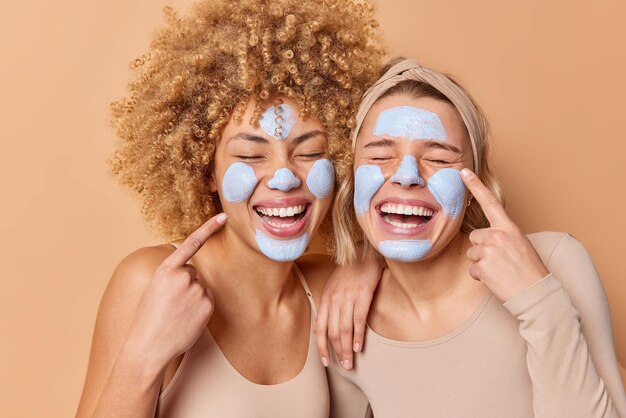 Les femmes ravies appliquent un masque d'argile de beauté pointent les doigts sur le visage se tiennent étroitement les uns aux autres gardent les yeux fermés subissent des soins du visage isolés sur fond beige Utilisez ce produit cosmétique