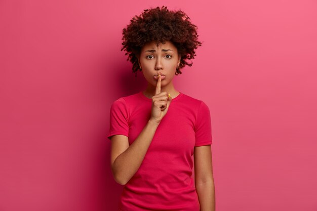 Ferme-la s'il te plaît. Déçu jeune femme afro-américaine fait un geste silencieux, montre des signes de silence, fronce les sourcils, appuie l'index sur les lèvres, porte un t-shirt décontracté, pose sur un mur rose