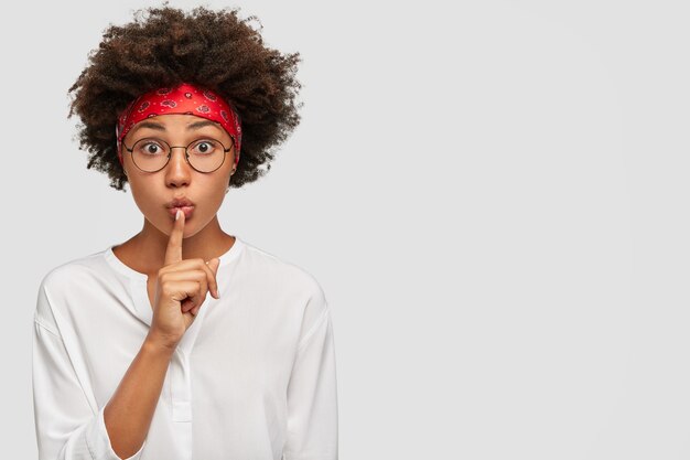 Ferme-la s'il te plaît! Une jeune fille sérieuse à la peau sombre garde l'index sur la bouche, montre un signe chut