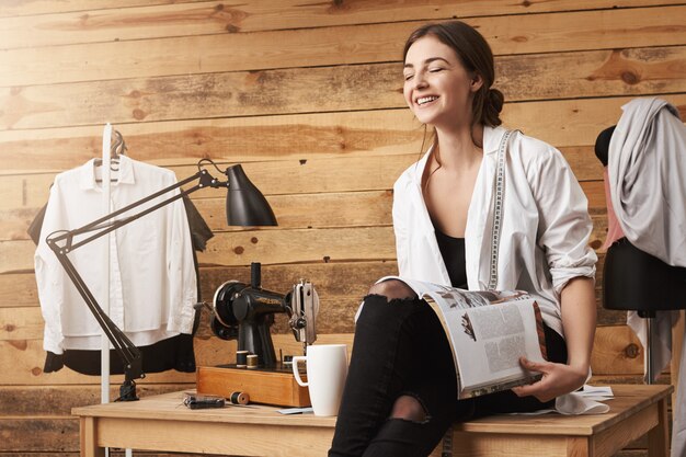 Fermez les yeux et voyez de nouvelles idées. Plan intérieur d'un égout heureux assis sur une table, lisant un magazine et buvant du thé, en pause, discutant des derniers potins avec un collègue. Fille et machine à coudre