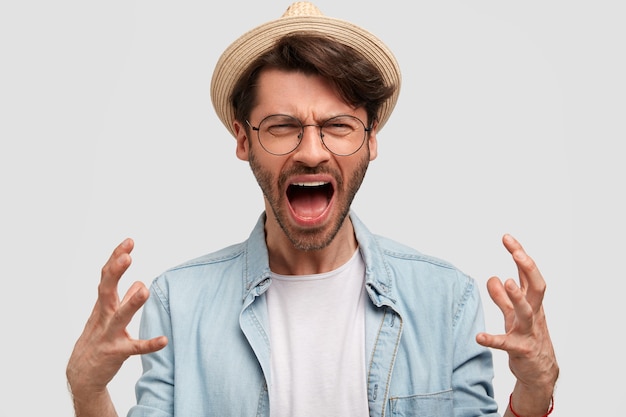 Fermier homme ennuyé avec chaume, gestes de colère et crie d'agacement, mécontent de la récolte, habillé en chapeau de paille et chemise en jean, pose contre le mur blanc