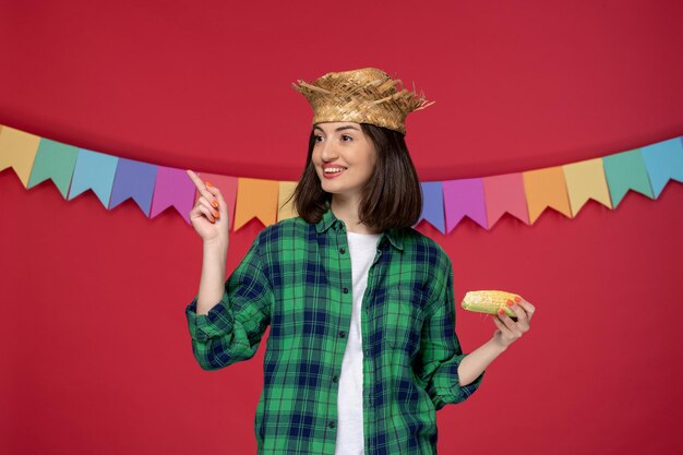 Festa junina jeune jolie fille en chemise verte célébrant le festival brésilien pointant avec du maïs