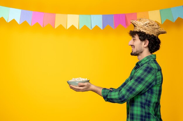 Photo gratuite festa junina jeune mec mignon en chapeau de paille et festival brésilien de drapeaux colorés donnant du pop-corn