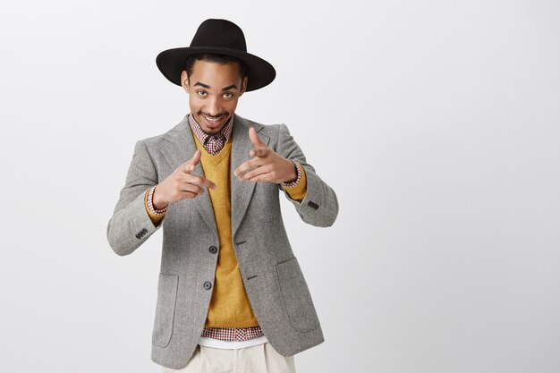 Un fêtard ému nous invitant au club. Portrait d'homme d'affaires beau positif dans un chapeau et une veste élégants, flirter ou être macho, pointant avec un geste de pistolet, debout sur un mur gris