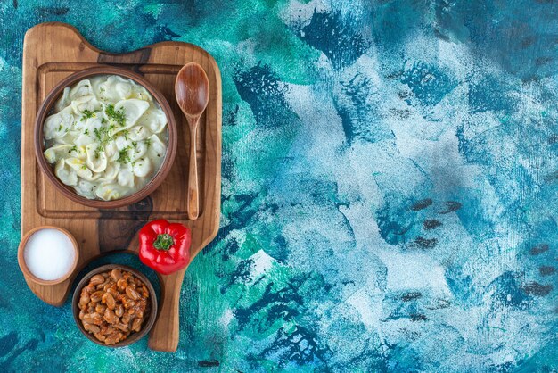 Fèves au lard, dushbara, cuillère, poivre et sel sur une planche, sur la table bleue.