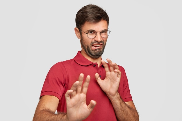 Photo gratuite fie! un homme barbu insatisfait repousse les mains, a une expression faciale dégoûtante, remarque quelque chose de détestable, fronce les sourcils avec aversion, porte un t-shirt rouge, isolé sur un mur blanc