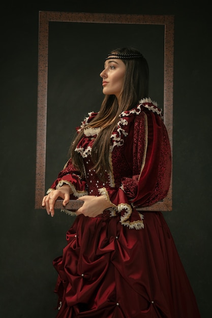 Fier. Portrait de jeune femme médiévale en vêtements vintage rouge debout sur fond sombre. Modèle féminin en tant que duchesse, personne royale. Concept de comparaison des époques, moderne, mode, beauté.