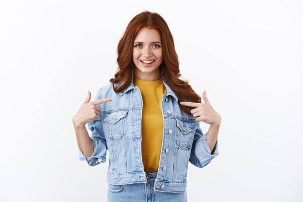 Fière et confiante, motivée, jolie fille au gingembre avec des taches de rousseur dans une veste en jean, se pointant du doigt et souriante vantarde, se sentant honorée et sûre d'elle, suggère de l'aide, atteint l'objectif, mur blanc