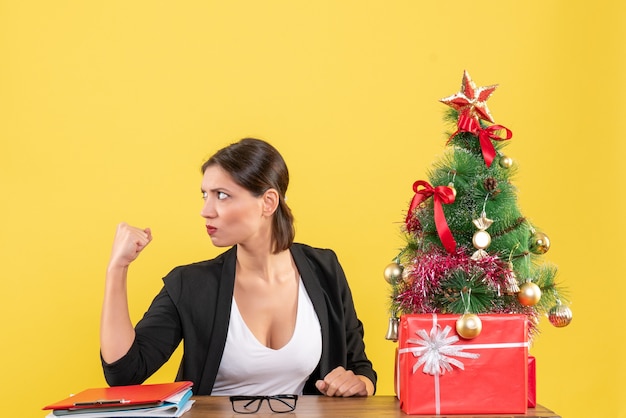 Fière jeune femme en costume avec arbre de Noël décoré au bureau sur jaune