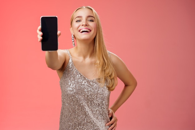 Fière joyeuse charmante femme européenne blonde gaie dans une élégante robe brillante argentée tenir la taille de la main confiante étendre le bras montrant l'affichage du smartphone présente un nouvel appareil d'application génial, fond rouge.
