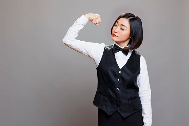 Photo gratuite une fière serveuse asiatique en uniforme professionnel montrant de forts muscles du biceps. une jeune réceptionniste attrayante montrant sa force et sa puissance tout en posant en studio sur un fond gris