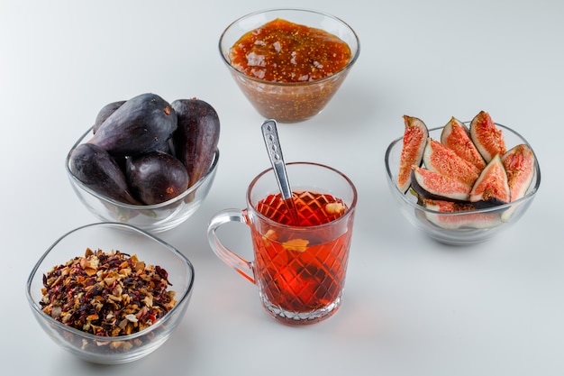 Figues avec confiture de figues, thé, cuillère à café, herbes séchées dans des bols sur blanc, high angle view.