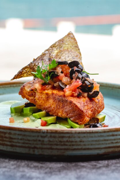 Filet de poisson frit placé sur des tranches d'avocat et une sauce garnie d'olive tomate et de peau de poisson