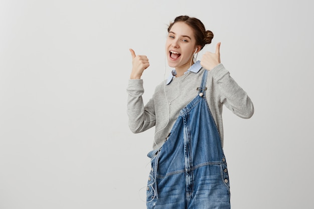 Photo gratuite fille de 20 ans en denim étant heureux gesticulant pouce en l'air sur le mur blanc. écrivain infantile femelle célébrant le succès de son nouveau livre en l'écoutant en version électronique via des écouteurs. concept technique