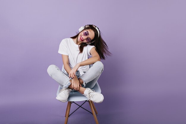 Fille adorable à lunettes de soleil assis sur une chaise avec les jambes pliées. Portrait de femme brune séduisante appréciant la musique.