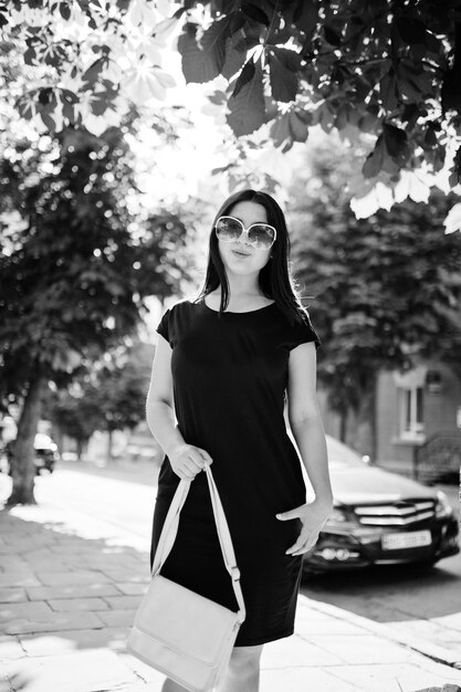 Fille d'affaires brune en robe noire sur des lunettes de soleil avec sac à main à la main posant dans la rue de la voiture de fond de la ville sur la route