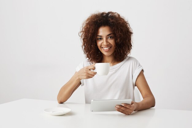 Fille africaine souriante tenant la tasse et la tablette assis à table sur le mur blanc.