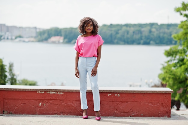 Une fille afro-américaine bouclée posée dans la rue de la ville porte un chemisier rose et un pantalon bleu