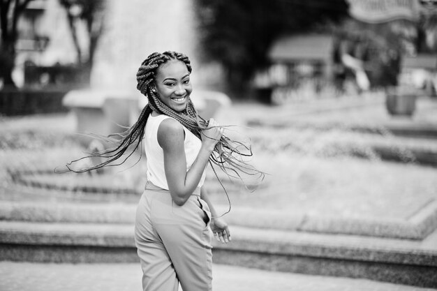 Fille afro-américaine à la mode au pantalon rose et aux dreads rouges posées en plein air contre les fontaines