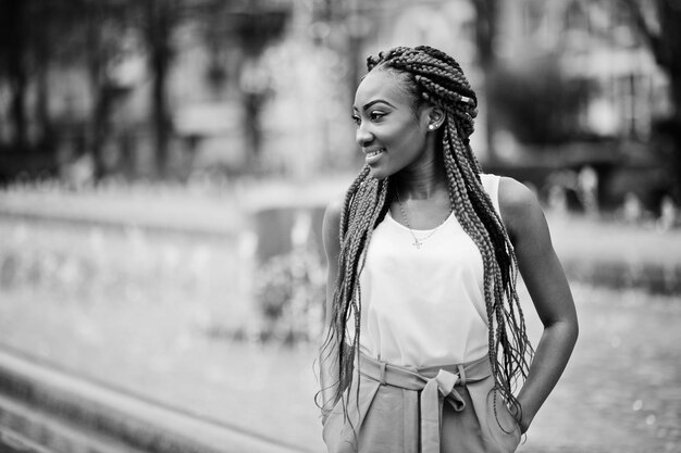 Fille afro-américaine à la mode au pantalon rose et aux dreads rouges posées en plein air contre les fontaines